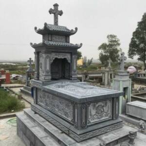 Mộ đá Công giáo hai mái bằng đá xanh, Thánh giá trên đỉnh mái, thân mộ chạm chữ IHS và dây nho, trong nghĩa trang giáo xứ